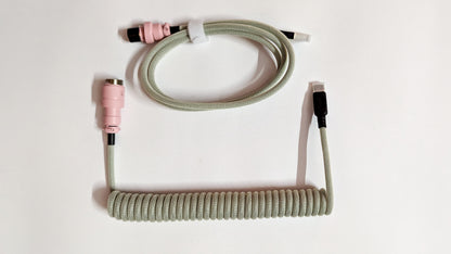 A coiled keyboard cable with a pink connector on one end and a USB-C connector on the other. The cable is gray and has a braided design.