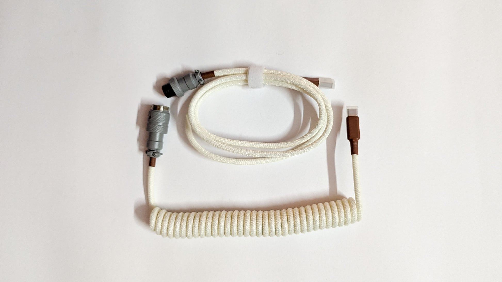 A coiled keyboard cable with a gray connector on one end and a USB-C connector on the other. The cable is white and has a braided design.